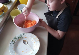 Chłopiec stojący przy blacie kuchennym, ubrany w fartuszek miesza utartą marchewkę w misce.